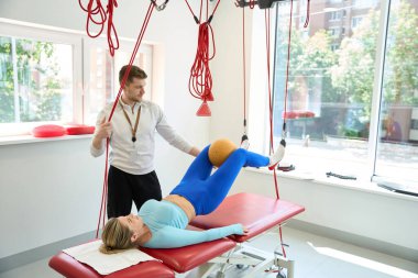 Bacaklarının arasında top olan güzel bir kadın içeride kırmızı kablo kullanarak antrenörle süspansiyon terapisi yapıyor.