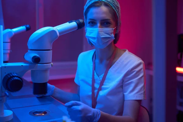 Laboratory assistant in a protective mask uses a powerful microscope, female works in protective gloves