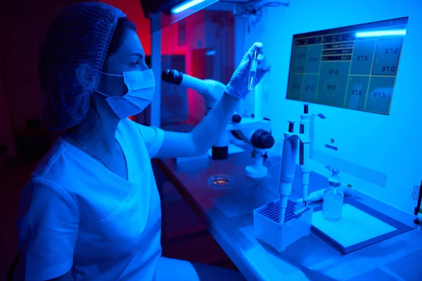 女性の胚科医は 実験室の強力な顕微鏡で 彼の手でバイオマテリアルを持つ試験管を保持しています — ストック写真