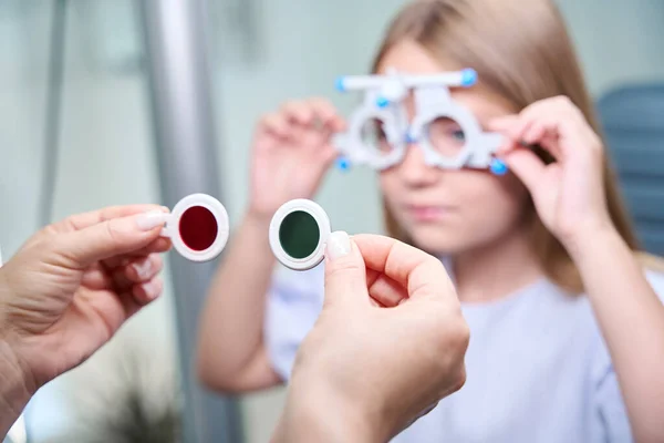 Ophthalmologist hands holding red and green filters in front of child with ophthalmic trial frame on face