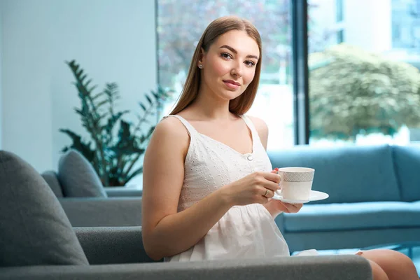 Young woman relaxing with a cup of tea on a cozy sofa, in the hospital foyer eco-friendly design