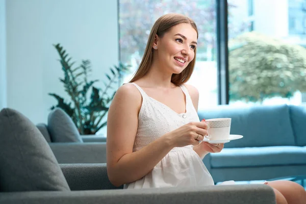 Cute young woman sitting with a cup of tea on a cozy sofa, in the hospital foyer eco-friendly design
