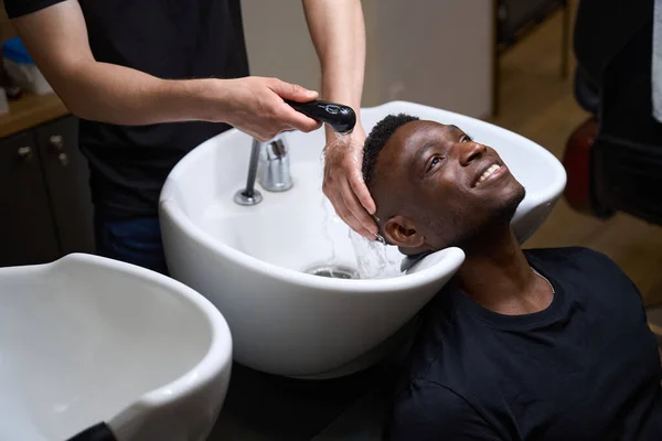 Barber washes a guys hair in a special sink, his African American client has curly hair