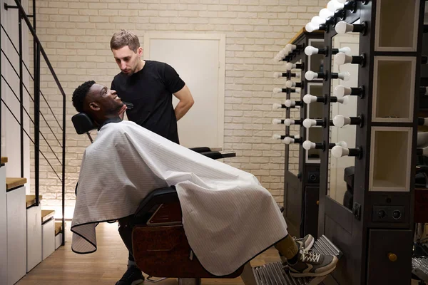 Stylist works in front of an illuminated mirror as he shaves an African American client with an electric razor