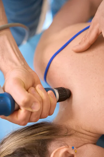 Male therapist affecting on trigger point on female shoulders using special device for extracorporeal shockwave therapy, close-up