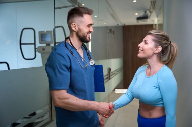 Minnettar kadın hasta, çabuk iyileşmesi için travma uzmanına el sıkışıyor. Tıp merkezinde rehabilitasyon programı var.