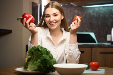 Kameraya bakıp içeride otururken elinde dolmalık biber ve domates tutan gülümseyen kadın.