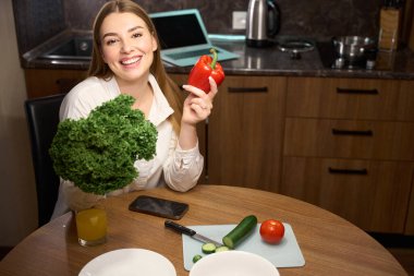 Kameraya bakıp evde otururken elinde çan biberi ve marul yaprakları tutan gülümseyen bir kadın.