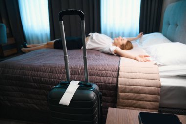 Otelde arka planda rahat bir yatakta uzanmış bir kadının olduğu bavul fotoğrafı.