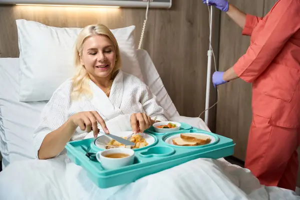 Woman in hospital on bed rest is having lunch in bed, next to a nurse preparing an IV for her
