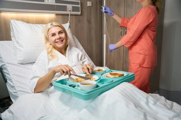 Recovering patient on bed rest is having lunch in bed, with a nurse nearby preparing an IV for her
