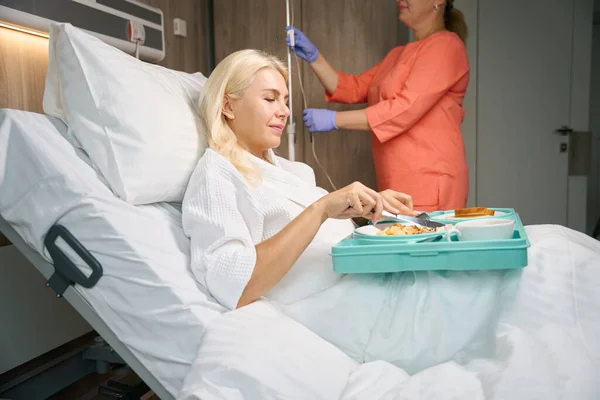 침대에서 여자를 회복하는 그녀를 Iv를 준비하는 간호사 침대에서 점심을 먹고있다 — 스톡 사진