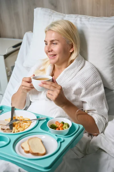 Recovering female patient in a hospital gown is having breakfast in bed, she is in hospital mode
