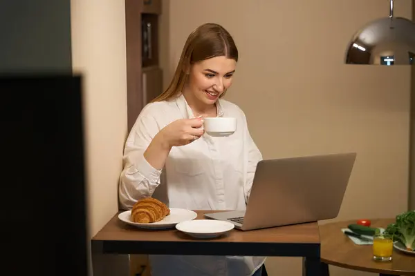 Smiling woman in casual clothes browsing laptop while snacking and standing in kitchen
