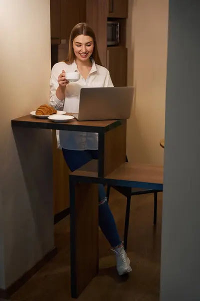 Happy woman in casual clothes browsing notebook computer while snacking and standing indoors