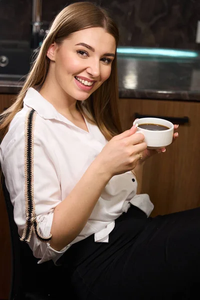 Smiling female in fancy clothes looking at camera while holding coffee cup and sitting on chair indoors