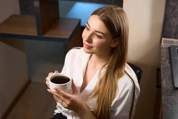 Pretty female in fancy clothes holding cup of coffee while sitting on chair indoors