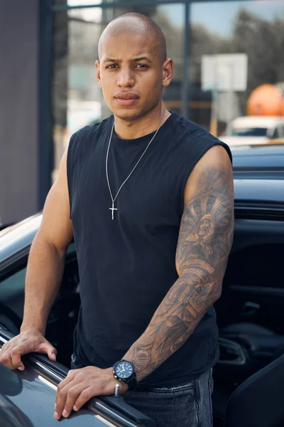 Handsome African American man with tattoo on hand standing near his luxury car and looking at camera, engaging in car transportation and delivery