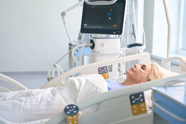 Ameliyattan sonra kadın sarışın hastaların hayati fonksiyonlarını izlemek için modern ekipmanlarla dolu bir odada yatıyor.