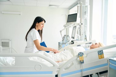 Bakıcı hemşire ameliyat sonrası hastayla iletişim kurar, elini tutar.