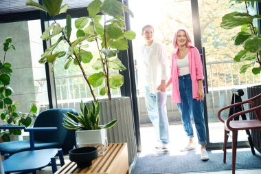 Gülümseyen genç adam ve kadın el ele tutuşuyor, bir tasarım stüdyosuna giriyor, odada bir sürü canlı bitki var.