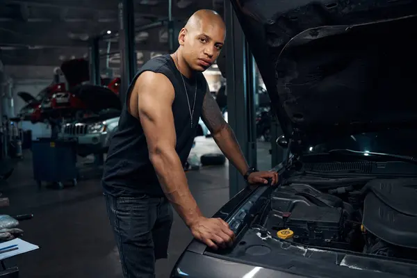 African American masculine man looking at camera with serious face expression standing near car with opened hood, going to make system diagnostic and some repairing works