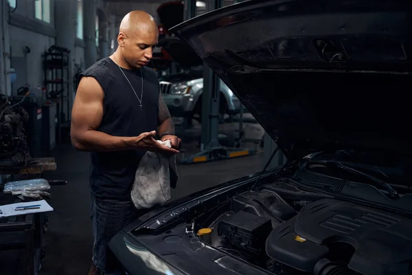 Serious repairman trying to clean his hands from motor oil and grease with rag, making pause while repairing luxury car, completing system diagnostic at vehicle service station