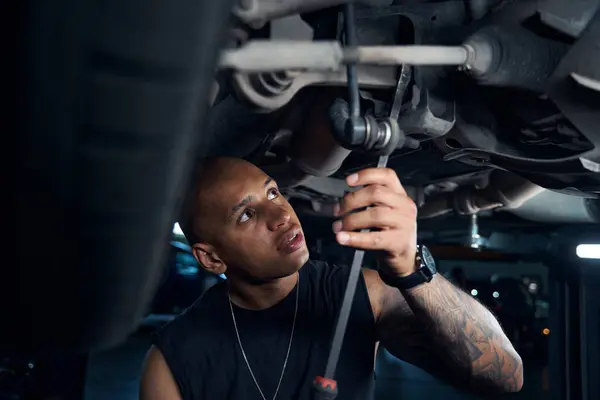 African American mechanic checking and repairing car axle, screwing spare parts with long screwdriver, adjusting or altering chassis standing under the car, auto service station