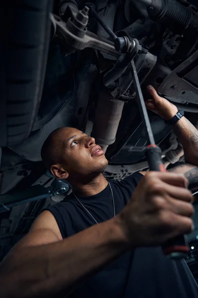 Focused African American car-mechanic checking car axle and lever integrity using long scrap, car bodywork, motor vehicle service station