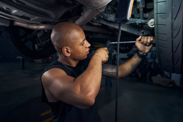 Serious African American repairman checking the condition and reliability of fastening of front suspension arms to the ball joint, repairing car in motor vehicle service station