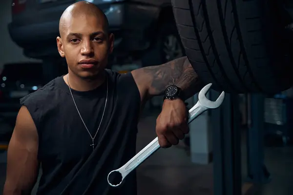 Handsome African American man auto-mechanic holding big socket wrench in hand, looking seriously, engaging in reconditioning and adjustment of any components of motor vehicle
