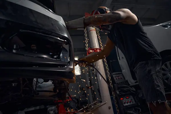 Masculine African American auto-mechanic welding rusty car body that hanging on car lift in his garage wearing protective gloves and face mask, automobile repair shop