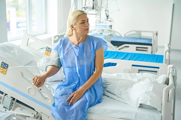 Woman in a hospital gown sits on bed in the recovery room, she is connected to support and monitoring machines