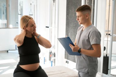 Hamile bir kadın boyun ağrısı hakkında fizyoterapiste şikayette bulunuyor. Her şeyi mavi bir klasöre yazıyor.