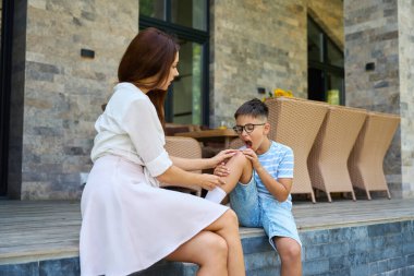 Kadın, oğullarının dizindeki yarayı alçıyla kapatıyor. Bir kır evinin terasında oturuyorlar.