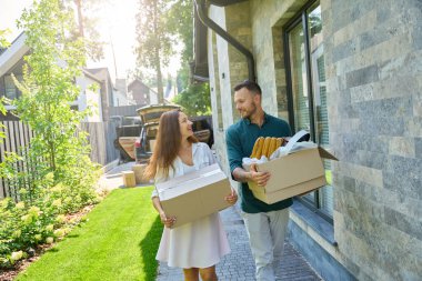 Karı koca açık bir bagajdan kutu taşıyorlar, bir kır evinin bahçesinde yürüyorlar.