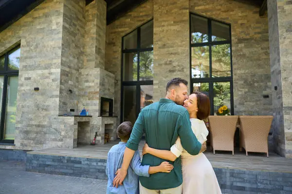 Sweet family stands hugging in front of a country house, dad kisses mom