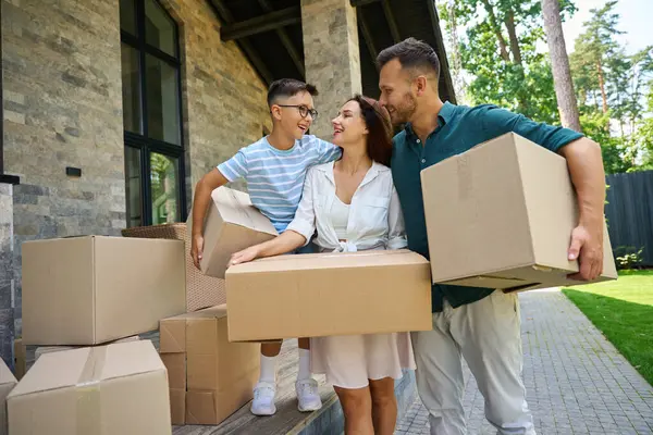 Young parents and their son move boxes of things to their new home, they chat cheerfully