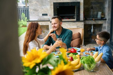 Dost canlısı aile, baba, anne ve oğul, terasta öğle yemeği yiyorlar, erkek kadına bir parça incelik gösteriyor.