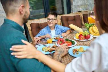 Anne ve baba öğle yemeğinde terasta oğluyla iletişim kurar. İnsanlar rahat bahçe mobilyalarını kullanır.