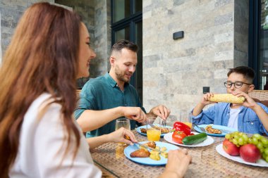 Oğlan iştahla mısır yiyor, aile bir kır evinin terasında öğle yemeği yiyor.