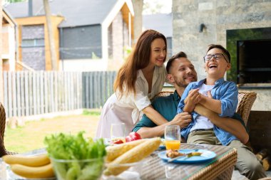 Çocuk yemek masasında neşeyle gülüyor, ailesi ona nazikçe sarılıyor.