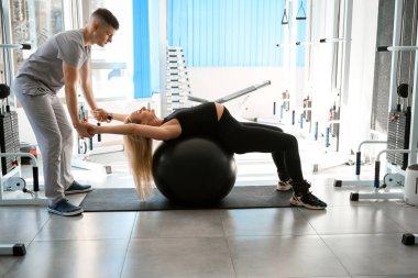 Hamile kadın özel egzersizleri bir fitball topuyla yapıyor, eğitmen ellerini tutuyor.