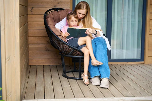可爱的金发母亲和她的可爱的女儿 有着时髦的发型 坐在木制门廊的扶手椅上看书 愉快地消遣着 — 图库照片