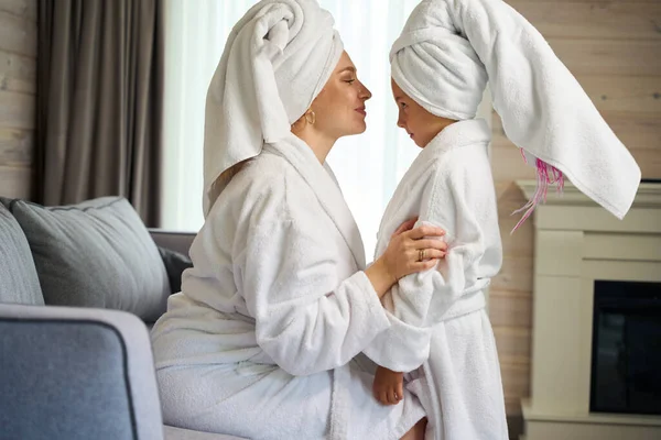 Mother and daughter enjoying pastime together in spa, standing in hotel room in soft bathrobes and big towels on heads, mother kissing her daughter