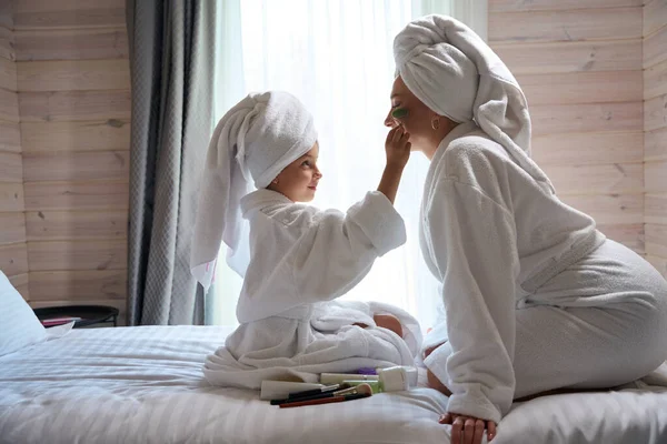 漂亮的小女孩穿着大浴衣 头上戴着毛巾 把化妆品涂在她母亲洁白的脸上 女孩们一起在温泉后很开心 — 图库照片