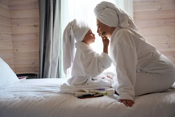穿着浴衣头戴浴巾的可爱小女孩在妈妈亲吻女儿的时候用特殊的工具温柔地按摩着妈妈的脸 — 图库照片