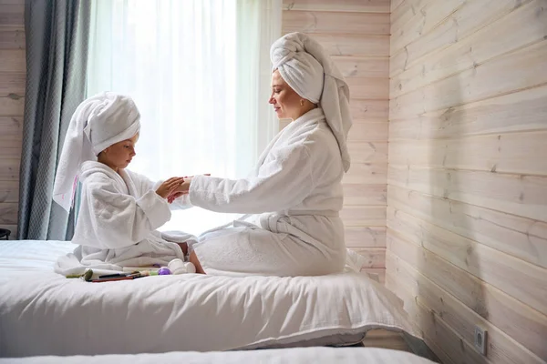 Mother watching her daughter applying body lotion on her hands, body care after bath and spa, pleasure pastime together