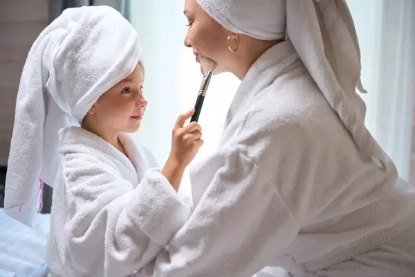 Pretty little daughter pretending she is cosmetician and applying blush on mother face after spa, girls having fun sitting in warm bathrobes