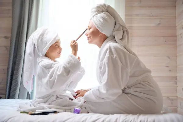 漂亮的小女孩给她美丽的母亲化妆 女孩们穿着浴衣坐在床上 头戴浴巾 美丽的一天 — 图库照片